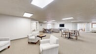Located at 500 College Ave in Terre Haute, Indiana, this single-floor community space offers a generous and well-appointed layout designed for social engagement and relaxation. The interior is characterized by neutral tones, plush carpeting, and an abundance of flush-mounted skylight panels that bathe each area in bright, even illumination throughout.

The expansive game room serves as a central hub, featuring multiple seating arrangements with upholstered armchairs and game tables ideal for chess and cards. Decorative framed artwork depicting card suit motifs adds a thematic touch to the walls. A mounted flat-screen television and comfortable sofa seating create a media lounge area within the same open space, while additional dining-style tables and chairs provide flexible gathering options.

The library area is a quiet, light-filled retreat highlighted by an elegant arched window that frames views of the surrounding greenery. Comfortable seating, a display case, and framed artwork depicting grand libraries lend the room a scholarly, relaxed ambiance.

A wide corridor connects the spaces and features arched windows, a textured ceiling, and restroom access, ensuring convenient circulation throughout. An additional lounge area with wainscoting, framed botanical prints, and coordinated upholstered seating rounds out this thoughtfully designed community space.