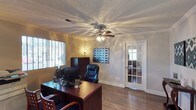 This single-floor space features a thoughtfully designed office room that combines functionality with classic architectural details. The room is finished with warm hardwood flooring that adds a sense of depth and character throughout. Crown molding lines the perimeter of the ceiling, lending a refined, polished look, while a textured ceiling adds subtle visual interest overhead. A ceiling fan with an integrated light fixture serves as the central focal point, providing both ventilation and ambient lighting.

Natural light filters in through a window fitted with horizontal blinds, offering control over brightness and privacy. The space is furnished with a rich, dark wood executive desk and matching cabinetry, complemented by a cushioned leather office chair that emphasizes a professional atmosphere. A decorative geometric artwork adorns one wall, adding a pop of visual interest.

A distinctive French door with glass pane inserts leads to an adjacent area, allowing borrowed light to pass through while maintaining a sense of separation between spaces. The overall ambiance is professional yet inviting, making this office well-suited for productive work in a comfortable, well-finished environment.