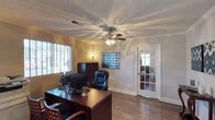 This single-floor space features a thoughtfully designed office room that combines functionality with classic architectural details. The room is finished with warm hardwood flooring that adds a sense of depth and character throughout. Crown molding lines the perimeter of the ceiling, lending a refined, polished look, while a textured ceiling adds subtle visual interest overhead. A ceiling fan with an integrated light fixture serves as the central focal point, providing both ventilation and ambient lighting.

Natural light filters in through a window fitted with horizontal blinds, offering control over brightness and privacy. The space is furnished with a rich, dark wood executive desk and matching cabinetry, complemented by a cushioned leather office chair that emphasizes a professional atmosphere. A decorative geometric artwork adorns one wall, adding a pop of visual interest.

A distinctive French door with glass pane inserts leads to an adjacent area, allowing borrowed light to pass through while maintaining a sense of separation between spaces. The overall ambiance is professional yet inviting, making this office well-suited for productive work in a comfortable, well-finished environment.
