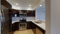 This single-floor space showcases a well-appointed kitchen that combines modern aesthetics with practical design. The kitchen is anchored by rich, dark espresso-toned cabinetry that runs both along the lower base and upper wall sections, providing ample storage throughout. The cabinetry's sleek, flat-panel doors with minimalist hardware contribute to the contemporary feel of the space.

Light-toned stone countertops offer a striking contrast against the dark cabinets, creating a visually balanced workspace. The kitchen is equipped with a full suite of stainless steel appliances, including a gas range, built-in microwave, and dishwasher, all seamlessly integrated into the layout.

The sink area is positioned along a peninsula-style countertop that opens toward an adjacent room, enhancing the sense of openness and connectivity within the space. Recessed lighting installed in the ceiling provides consistent, even illumination throughout the kitchen. The light-colored flooring and pale blue-grey walls further brighten the space, complementing the contrast of the dark cabinetry and creating a clean, cohesive interior atmosphere.