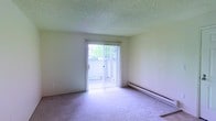 This single-floor space features a simply designed living area finished with neutral-toned wall-to-wall carpeting that lends a clean, understated aesthetic throughout. The room is illuminated by natural light streaming through a full-width sliding glass door fitted with vertical blinds, which can be adjusted to control light levels and privacy. The sliding door provides direct access to an outdoor area, where glimpses of surrounding greenery are visible, adding a touch of nature to the interior atmosphere. A baseboard heater runs along the exterior wall beneath the sliding door, offering a practical heating solution integrated seamlessly into the room's layout. The ceiling features a textured popcorn finish, consistent with the overall straightforward design of the space. The walls are painted in a soft, light tone, contributing to the room's bright and open feel. The layout is open and unobstructed, providing flexibility for various furniture arrangements. Overall, the space presents a functional and practical environment suitable for everyday living, with its primary highlights being the natural light, outdoor access, and understated finishes.