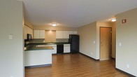 This single-floor unit features an open-concept layout that seamlessly connects the kitchen and living area, creating a functional and spacious feel. The kitchen is well-equipped with ample white upper cabinetry offering generous storage, dark countertops, and a full suite of black appliances including a refrigerator, dishwasher, and built-in microwave. A peninsula with a dark countertop extends from the kitchen, providing additional workspace or casual dining space. The sink is positioned along the back counter beneath the cabinetry, maximizing the efficiency of the kitchen layout. Warm hardwood-style flooring runs throughout the open living area, adding a cohesive and inviting aesthetic to the space. The neutral beige walls provide a clean, versatile backdrop suitable for a variety of interior styles. Flush-mount ceiling lighting illuminates the kitchen area effectively. A door visible on the far wall leads to an additional room or entry point, adding to the unit's overall connectivity. The layout is straightforward and practical, making efficient use of the available space on the single floor.