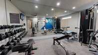 Located at 505 W Baseline Rd in Tempe, Arizona, this fitness center offers a well-equipped, single-floor workout facility designed to support a wide range of training styles. The space is defined by its open layout, wall-to-wall mirrors that visually expand the room and allow users to monitor their form, and recessed lighting that keeps the environment bright and functional throughout.

The gym is outfitted with a comprehensive selection of strength training equipment, including a Smith machine loaded with weight plates, a cable pulley machine ideal for a variety of resistance exercises, multiple adjustable benches suited for incline, flat, and decline positions, and a full rack of dumbbells organized on tiered storage racks. A kettle bell and step platforms round out the free weight area, providing versatility for functional training routines.

A bold geometric mural in blue, white, and teal anchors one wall, adding a dynamic visual element to the otherwise clean, neutral aesthetic. High-mounted windows along one wall allow natural light to filter in, complementing the recessed ceiling fixtures. A wall-mounted flat-screen television is positioned for easy viewing during workouts. The durable, textured flooring throughout the space provides stability and safety underfoot. The facility also includes clearly marked exit access, ensuring a safe and compliant environment for all users.
