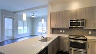This single-floor space showcases a thoughtfully designed open-concept layout that seamlessly connects the kitchen and living area. The kitchen is outfitted with stainless steel appliances, including a gas range, built-in microwave, and dishwasher. Light wood-toned cabinetry with sleek bar-pull hardware lines the walls, complemented by a textured tile backsplash that adds subtle visual interest. A generous island with a crisp white quartz countertop serves as the centerpiece of the kitchen, featuring a stainless steel undermount sink with a modern gooseneck faucet — ideal for both meal prep and casual dining.

Pendant lighting hangs above the island, drawing the eye toward the adjoining living area, which benefits from multiple windows that flood the space with natural light. A ceiling fan in the living area contributes to both the aesthetic and functional comfort of the room. Dark hardwood-style flooring grounds the space, creating a striking contrast against the white walls and light cabinetry. The overall ambiance is clean, contemporary, and welcoming, with a cohesive design language carried throughout.