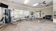 Located at 2201 Williams Glen Blvd in Zionsville, Indiana, this fitness center offers a thoughtfully designed, single-floor layout divided into two dedicated exercise rooms. The main workout area is bright and spacious, featuring large windows with blinds that allow generous natural light to fill the room while providing views of the surrounding greenery. Crown molding and neutral gray walls give the space a clean, polished aesthetic, while patterned carpet tiles in beige and teal add a contemporary touch underfoot. A ceiling fan overhead ensures comfortable air circulation during workouts. The main room is outfitted with a range of Cybex strength training machines, a cable pulley system, an adjustable weight bench, and a full rack of dumbbells. Cardio equipment includes treadmills, an elliptical, and a stationary bike, all positioned to take advantage of the natural light. A large wall-mounted mirror enhances the sense of space and supports proper form during exercise. French doors connect the main area to a secondary exercise room, which features additional equipment, stability balls, and a desk area. Posted fitness center rules ensure a structured and safe environment for all users. Colorful artwork accents the walls, adding a welcoming, vibrant energy to this well-appointed community fitness facility.