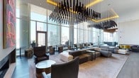 This striking single-floor space showcases an exceptional blend of contemporary design and refined comfort. The most commanding feature is its dramatic ceiling installation — a cascading arrangement of elongated black pendant fixtures suspended at varying heights, set against warm amber accent lighting that creates a bold visual focal point overhead.

The space benefits from an abundance of natural light, courtesy of expansive floor-to-ceiling glass windows that wrap around the perimeter, framing panoramic views of a dense urban skyline. The transparency of the glazing creates a seamless connection between the interior and the cityscape beyond.

The layout is thoughtfully arranged into distinct seating zones, accommodating both intimate conversations and larger gatherings. Furnishings include rich dark leather armchairs, modular sofas in neutral tones, and accent chairs upholstered in patterned fabric, adding visual interest throughout. Sleek coffee tables and side tables in glass and metal complement the modern aesthetic.

The flooring is finished in a light, smooth material that reflects the natural light, enhancing the overall sense of openness. Together, the architectural elements and curated furnishings create a polished, upscale ambiance suited to a high-end lounge or hospitality setting.