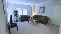 This single-floor space offers a welcoming and functional layout designed for everyday comfort. The living room serves as the focal point of the home, featuring plush carpeted flooring that adds warmth underfoot. Two generously sized dark upholstered sofas are arranged around a warm-toned wooden coffee table, creating an inviting seating area ideal for relaxing or entertaining. Crown molding lines the ceiling, lending the room a refined, classic character. Natural light filters through two windows dressed with white blinds, brightening the space with a clean, airy feel. Built-in shelving adds a practical yet decorative element along one wall, providing display and storage options. A potted indoor plant nestled in the corner introduces a touch of greenery and life to the room. A table lamp on a side table contributes soft ambient lighting, while a framed piece of wall art adds a decorative accent. The overall ambiance is calm, neutral, and versatile, with a light color palette across the walls and flooring that makes the space feel open and easy to personalize. The thoughtful arrangement and classic finishing details make this a comfortable, move-in-ready living environment.