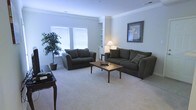 This single-floor space offers a welcoming and functional layout designed for everyday comfort. The living room serves as the focal point of the home, featuring plush carpeted flooring that adds warmth underfoot. Two generously sized dark upholstered sofas are arranged around a warm-toned wooden coffee table, creating an inviting seating area ideal for relaxing or entertaining. Crown molding lines the ceiling, lending the room a refined, classic character. Natural light filters through two windows dressed with white blinds, brightening the space with a clean, airy feel. Built-in shelving adds a practical yet decorative element along one wall, providing display and storage options. A potted indoor plant nestled in the corner introduces a touch of greenery and life to the room. A table lamp on a side table contributes soft ambient lighting, while a framed piece of wall art adds a decorative accent. The overall ambiance is calm, neutral, and versatile, with a light color palette across the walls and flooring that makes the space feel open and easy to personalize. The thoughtful arrangement and classic finishing details make this a comfortable, move-in-ready living environment.