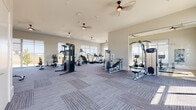 This single-level fitness center is designed to support a comprehensive range of workout routines in a bright, open environment. The space is divided into distinct functional zones, each thoughtfully outfitted with professional-grade equipment. The main workout floor features patterned carpet tile throughout and is lined with large windows that flood the area with natural light and provide views of the surrounding outdoor landscape, including a pool area visible from select vantage points.

The strength and cardio zone houses cable machines, weight benches, stair climbers, treadmills, and a variety of resistance machines, all arranged with ample spacing for comfortable use. Ceiling fans overhead contribute to a comfortable training environment, while full-length mirrors along one wall enhance the sense of space and allow for form monitoring.

A dedicated spin and functional training studio features light hardwood-style flooring, floor-to-ceiling windows with roller shades, and a row of smart stationary bikes. Battle ropes, TRX straps, a rack of free weights, and a wall-mounted display round out this versatile room.

The facility is accessed via a bright, gallery-style corridor with large-format tile flooring, framed artwork, and views into adjacent amenity spaces. The overall aesthetic is clean, modern, and purposeful, creating an inviting atmosphere for daily fitness routines.