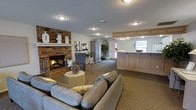 This inviting single-floor space combines warmth and functionality in a well-appointed layout designed for comfort and community. The lounge area serves as the heart of the space, anchored by a classic full-height brick fireplace that adds a rustic, homey character. Plush, neutral-toned sofas and accent chairs are arranged to encourage relaxed conversation, complemented by patterned carpeting that ties the seating area together. Decorative accents on the fireplace mantel and surrounding surfaces add thoughtful styling throughout.

Adjacent to the lounge, a built-in reception or service counter features light wood cabinetry with upper and lower storage, creating a practical and organized workstation. A window above the counter allows natural light to filter into the space, brightening the area and offering a connection to the outdoors.

Recessed flush-mount ceiling fixtures provide consistent, warm overhead lighting throughout. A tall armoire and potted greenery add vertical interest and a touch of nature to the interior. A glass door at the rear of the space suggests easy access to an outdoor area, further enhancing the open and accessible feel. Overall, the space strikes a balance between a residential warmth and a functional communal environment.