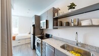 Located at 120 20th Ave S in Nashville's vibrant Midtown neighborhood, this efficiently designed one-bedroom, one-bathroom unit makes smart use of its compact footprint across a single floor. The layout includes a bedroom, kitchen, bathroom, and hallway, each thoughtfully appointed with contemporary finishes throughout.

The bedroom is a bright, well-organized space featuring large windows with horizontal blinds that frame views of the surrounding Nashville cityscape, allowing generous natural light to fill the room. Light wood-tone flooring runs throughout, complementing the clean white walls. The room accommodates a full bed with warm rust-orange bedding, a wall-mounted flat-screen TV, a dedicated desk area with a task chair, and a full-length mirror.

The kitchen is modern and functional, outfitted with slate-gray cabinetry, granite countertops, a stainless steel sink with a pull-down faucet, and stainless steel appliances including a range and microwave. Open floating shelves above provide additional storage and display space.

The bathroom continues the clean aesthetic with white walls, wood-look flooring, and a dark-paneled door that adds contrast. The space is straightforward and well-maintained. Overall, the unit offers a cohesive, modern design well-suited for urban living in the heart of Nashville.