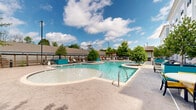 Located at 6854 Governors West NW in Huntsville, Alabama, this outdoor amenity space showcases a generously sized, freeform swimming pool as its central feature. The pool's curving, organic shape is surrounded by a spacious stamped concrete deck, designed to accommodate a variety of leisure activities. Mature trees are thoughtfully positioned throughout the deck, providing natural shade and greenery that enhances the resort-like atmosphere.

The lounge areas are well-appointed with modern reclining chairs and accent tables, offering comfortable spots for relaxation both poolside and near the privacy fencing. The horizontal-slat wooden fencing adds a contemporary, stylish boundary to the space while maintaining an open, airy feel. A safety ring and equipment station are also present, underscoring the space's commitment to safety.

Vibrant teal accent features, including planter walls and pool furnishings, complement the clear blue water and bright sky, contributing to a lively, welcoming ambiance. Shaded seating areas with umbrellas provide additional comfort along the pool's perimeter. The surrounding multi-story building with balconies frames the outdoor area, seamlessly connecting indoor and outdoor living. Overall, this meticulously designed pool area delivers a dynamic, community-centered outdoor experience.