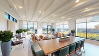 This thoughtfully designed single-floor amenity space seamlessly blends a formal meeting area with a relaxed social lounge, creating a versatile environment suited for both productive gatherings and casual interaction. The architecture is defined by striking exposed beam ceilings with recessed lighting, crisp white walls, and large-format floor-to-ceiling windows that flood the interior with natural light and frame sweeping outdoor views of the surrounding landscape and rooftop terrace.

The conference and dining zone anchors one end of the space, centered around a substantial natural wood table paired with teal upholstered chairs atop a bold geometric black-and-white patterned floor. A wall-mounted large-screen display and a sleek built-in credenza with a textured, glimmering accent wall behind it provide both functionality and visual interest. A beverage station and additional storage complete this area.

The lounge area flows organically from the dining zone, furnished with retro-inspired striped armchairs in warm amber tones, wire-frame accent chairs, and cylindrical side tables that add an earthy warmth. Lush potted topiaries and greenery are strategically placed throughout, enhancing the fresh, airy ambiance. Roller shades offer flexible light control while preserving the connection to the bright, verdant outdoor views.