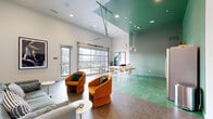 Located at 757 N Grant St in Denver, CO, this single-floor space offers a thoughtfully designed open-concept layout that seamlessly integrates a kitchen, dining area, and living room. The space is defined by its striking use of color and contemporary furnishings, creating a lively yet functional environment.

The kitchen features light maple cabinetry, teal-toned countertops and backsplash, and stainless steel appliances including a full-size refrigerator, dishwasher, and microwave. The vibrant green epoxy flooring grounds this zone and extends into the dining area, adding a bold visual anchor to the space.

The dining area centers around a long communal-style table with bar-height stools, positioned beside a large glass garage-style door that opens to an outdoor terrace, flooding the area with natural light and offering views of the Denver skyline. This indoor-outdoor connection is a standout feature of the space.

The living area transitions to a polished concrete floor and boasts soaring ceilings that amplify the sense of openness. A wall-mounted large-screen television anchors the space, complemented by a teal sofa, a pair of rounded amber accent chairs, and curated artwork on the walls. Recessed lighting throughout ensures a clean, modern aesthetic. The overall ambiance is energetic, contemporary, and well-appointed.