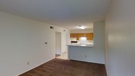 This single-floor unit presents an open-concept layout that connects the living area to the kitchen, creating a functional and cohesive living environment. The living area is finished with wall-to-wall brown carpet, providing a warm underfoot feel, while neutral-toned walls offer a versatile backdrop throughout the space.

The kitchen is positioned at the back of the open area and is accessible via a peninsula counter that doubles as a breakfast bar or workspace. Warm honey-toned wood cabinetry lines the upper walls of the kitchen, offering ample storage. The kitchen is equipped with a built-in range and a countertop sink, with under-cabinet lighting illuminating the work surfaces. The flooring in the kitchen transitions to a lighter, hard-surface material, distinguishing it from the carpeted living space.

A doorway visible from the living area leads to an adjacent room, with a louvered closet door nearby suggesting convenient storage access. The unit includes ceiling-mounted ventilation and is lit efficiently with overhead fluorescent fixtures in the kitchen. The overall ambiance is clean, simple, and practical.