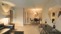 This single-floor residence exudes a warm, welcoming ambiance through its thoughtfully designed interior. The space is characterized by a neutral color palette of soft beige and cream tones that flow consistently throughout, creating a cohesive and calming atmosphere.

The dining area serves as a focal point of the living space, featuring a dark wood dining table paired with matching upholstered chairs. A distinctive circular pendant light fixture with a warm glow hangs above, complemented by a round decorative mirror on the wall that adds depth and visual interest. An elegant floor vase with tall natural branches contributes an organic, artistic touch to the corner of the room.

The floors are covered in plush, light-toned carpeting that adds comfort and warmth underfoot. A console table with sleek metallic legs anchors one side of the space, styled with decorative accents including candle holders.

The layout is open and connected, with a hallway visible in the background that leads to additional rooms, hinting at the functional flow of the floor plan. The smooth, light-colored walls and clean white baseboards contribute to a polished, well-maintained aesthetic. Overall, the space strikes a balance between contemporary style and comfortable livability, making it both functional and visually appealing.