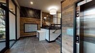 This contemporary residential lobby is thoughtfully designed to make a strong first impression. The entry is defined by large-format slate-toned tile flooring that extends throughout the space, grounding the design with an industrial yet refined aesthetic. Reclaimed wood planks clad the accent walls, adding warmth and texture that contrasts beautifully with the dark, matte black framing details and trim used throughout.

A striking geometric mural rendered in white line work on a dark background serves as a dramatic focal point, rising toward the ceiling and reinforcing the building's modern, urban character. The lobby features a built-in mailroom with a full array of individual resident mailboxes, set into the reclaimed wood wall for a seamless, integrated look. A package drop station with a clean white cabinet base provides additional mail-handling functionality.

The space is split-level, with a staircase featuring black metal railings leading to an upper lounge area furnished with seating and accent lighting. A stainless steel elevator provides accessible vertical circulation. Floor-to-ceiling glass entry doors and large windows flood the space with natural light while offering views of the surrounding greenery. A secondary corridor leads to additional building amenities, accented by a bold orange feature wall that adds a vibrant pop of color to the otherwise neutral palette.