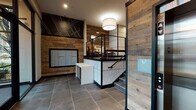 This contemporary residential lobby is thoughtfully designed to make a strong first impression. The entry is defined by large-format slate-toned tile flooring that extends throughout the space, grounding the design with an industrial yet refined aesthetic. Reclaimed wood planks clad the accent walls, adding warmth and texture that contrasts beautifully with the dark, matte black framing details and trim used throughout.

A striking geometric mural rendered in white line work on a dark background serves as a dramatic focal point, rising toward the ceiling and reinforcing the building's modern, urban character. The lobby features a built-in mailroom with a full array of individual resident mailboxes, set into the reclaimed wood wall for a seamless, integrated look. A package drop station with a clean white cabinet base provides additional mail-handling functionality.

The space is split-level, with a staircase featuring black metal railings leading to an upper lounge area furnished with seating and accent lighting. A stainless steel elevator provides accessible vertical circulation. Floor-to-ceiling glass entry doors and large windows flood the space with natural light while offering views of the surrounding greenery. A secondary corridor leads to additional building amenities, accented by a bold orange feature wall that adds a vibrant pop of color to the otherwise neutral palette.