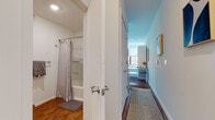 Located at 253 Academy St in Jersey City, NJ, this single-floor unit offers a clean, modern aesthetic throughout. Warm hardwood flooring runs continuously across the space, creating a cohesive and inviting atmosphere from room to room.

The kitchen is designed with functionality in mind, featuring bright white cabinetry extending from floor to ceiling, providing ample storage. Light countertops complement the white appliances, which include a range, microwave, refrigerator, and a compact dishwasher. A stainless steel sink with a modern faucet anchors the counter space, making the kitchen practical for everyday use.

The living area is compact yet well-organized, benefiting from a large window that allows natural light to fill the room and offers views of the surrounding neighborhood. A wall-mounted PTAC unit provides climate control, and colorful artwork adds personality to the otherwise neutral palette.

The bathroom includes a tub-shower combination with a curtain rod, a vanity with an integrated sink, a mirrored medicine cabinet, and clean white finishes throughout. A hallway connects the bathroom to a secondary vanity area, providing additional grooming space.

Overall, the unit maintains a minimalist, well-lit ambiance with thoughtful use of space.