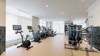 Located at 46 John St in Stamford, Connecticut, this fitness center offers a thoughtfully designed exercise space across a single floor. The room is finished with a clean, modern aesthetic, featuring white walls, a dropped ceiling with recessed lighting, and durable carpet flooring throughout.

The space is generously equipped to support a full range of workout routines. A cardio zone lines one side of the room, housing multiple treadmills, elliptical machines, stationary bikes, and an assault bike, several of which are equipped with personal screens. Opposite, a strength training area features a variety of cable and resistance machines, including leg press and multi-cable systems, alongside a power rack complete with a punching bag and TRX suspension trainers. A dedicated free weights section offers a full dumbbell rack, adjustable benches, and additional accessories.

Large windows along the perimeter allow natural light to fill the space, contributing to an open and energizing atmosphere. Full-length mirrors are strategically placed to enhance the sense of space and support proper form during training. Tiled accent walls near the entrance add a refined touch, framed by warm-toned wood-framed glass doors. A wall-mounted screen provides an additional functional element. The overall environment is clean, well-organized, and purpose-built for serious fitness use.