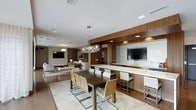This contemporary single-floor space showcases a thoughtfully designed open-plan layout that seamlessly integrates the dining, kitchen, and living areas into one cohesive environment. The interior is defined by a refined palette of warm walnut wood paneling, crisp white surfaces, and neutral tones that create an inviting yet sophisticated ambiance throughout.

The dining area takes center stage with a dark wood rectangular table surrounded by sleek chrome-framed chairs with natural wood backs, all positioned atop a light-toned area rug. Overhead, a striking sculptural chandelier with an organic branch-like design serves as a compelling focal point.

Adjacent to the dining space, the kitchen features a generous light stone countertop island with integrated bar seating and white upholstered stools. Wood cabinetry with recessed panel lighting and a wall-mounted flat-screen television enhance the functionality and style of this zone.

The living area beyond offers a comfortable lounge arrangement with upholstered seating, a wall-mounted television, and subtle ambient lighting. Recessed ceiling lights are distributed throughout, ensuring even, well-considered illumination across the entire floor plan. The cohesive design language and high-quality finishes make this a polished and functional space.