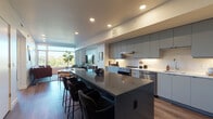This contemporary single-floor residence is thoughtfully designed with a clean, modern aesthetic throughout. The open-concept kitchen and living area serves as the heart of the home, anchored by a substantial kitchen island with a dark stone countertop and seating for multiple guests. The kitchen is outfitted with flat-front gray cabinetry, a white subway tile backsplash, stainless steel appliances, and under-cabinet lighting that enhances the bright, functional workspace. Wide-plank hardwood flooring flows seamlessly through the main living areas, adding warmth to the contemporary design. The living room is bathed in natural light from floor-to-ceiling windows that frame lush treetop views, creating a serene and airy atmosphere. The primary bedroom continues the connection to the outdoors with large windows overlooking the tree canopy, while soft carpeting adds comfort underfoot. A well-organized walk-in closet with adjustable wire shelving is conveniently located adjacent to a tiled bathroom featuring a glass-enclosed shower. A secondary bedroom provides additional flexible living space. The dedicated laundry room includes a full-size side-by-side washer and dryer with overhead shelving for added storage. Recessed lighting throughout the residence ensures a consistently polished and well-lit environment.