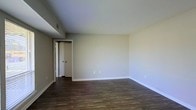 This single-floor space presents a straightforward and functional interior highlighted by rich, dark hardwood flooring that extends throughout the main living area. The floors provide a warm contrast against the neutral, light gray-beige walls and crisp white baseboards, creating a clean and cohesive aesthetic. The ceiling is smooth and painted white, contributing to a bright, open atmosphere within the room.

The space is unfurnished, allowing for a clear view of the room's proportions, which offer ample space for a variety of furniture arrangements. A doorway visible from the main area leads to an adjoining room or hallway, suggesting a practical and connected floor plan. The white-paneled interior door features classic round hardware, maintaining a simple and timeless design throughout.

A ceiling vent is visible, indicating a central HVAC system for climate control. Electrical outlets are positioned along the lower walls at standard heights, providing convenient access throughout the space. The overall ambiance is calm and neutral, offering a versatile blank canvas well-suited for personalization. The combination of durable flooring and muted wall tones gives the space a polished yet understated character.
