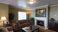 This single-floor home exudes a warm, classic charm throughout its thoughtfully designed living spaces. The living room serves as the heart of the home, anchored by a elegant fireplace with a crisp white mantle and neutral tile surround — an ideal focal point for the room. Rich crown molding frames the textured ceiling, adding a refined architectural detail that elevates the overall aesthetic. Warm olive-toned walls create an inviting and cozy atmosphere, complemented by a plush, earth-toned sofa and a traditional area rug that together contribute to the room's welcoming character. A dark wood coffee table and matching console table provide functional yet stylish accents. Natural light filters in through a large window dressed with horizontal blinds, brightening the space while maintaining privacy. A framed landscape painting above the fireplace adds a touch of artistic character. The overall ambiance is one of comfortable, traditional living — a space that balances everyday practicality with timeless design sensibilities, making it an ideal setting for both quiet relaxation and casual entertaining.
