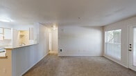 Located in Beaverton, Oregon, this one-bedroom, one-bathroom unit offers a well-organized layout across a single floor. The combined living and dining area is bright and open, featuring neutral-toned carpet, light walls, and a wall-mounted heating unit. A sliding glass door with an adjacent window floods the space with natural light and provides direct access to a private balcony with outdoor views. The kitchen is positioned adjacent to the living area and features a peninsula counter with a built-in sink, white cabinetry with light wood trim, a full-size refrigerator, and an electric range — all under bright overhead lighting. The bedroom is a comfortable, carpeted room with a window that brings in natural light and includes baseboard heating. Three closets throughout the unit provide generous storage options. The bathroom includes a vanity with under-sink cabinetry, a large mirror, wood-style flooring, and a combined tub and shower. A dedicated laundry area and a hallway entrance complete the layout. The unit maintains a clean, neutral aesthetic throughout, with consistent light-toned finishes across all rooms.