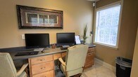 This single-floor space offers a thoughtfully designed environment with a focus on functionality and comfort. The home office area is a standout feature, boasting a built-in wooden desk with warm cabinetry and ample drawer storage, providing generous workspace for productivity. Dual computer monitors are positioned side by side, making the setup ideal for multitasking or collaborative work. A cushioned rolling office chair in a neutral tone anchors the workstation, offering both comfort and style. Natural light streams in through a large window dressed with white horizontal blinds, creating a bright and inviting atmosphere. The walls are finished in warm, earthy tones that complement the honey-toned wood cabinetry and light tile flooring throughout. A decorative framed artwork with an ornate gold frame adds a touch of elegance to the wall above the desk. A tall decorative plant in the corner introduces a natural, refreshing element to the room. The overall ambiance is warm, organized, and professional, making this space well-suited for focused work or administrative tasks in a comfortable, home-like setting.