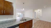 This 2-bedroom, 2-bathroom residence offers a practical and well-designed single-floor layout with a clean, modern aesthetic throughout. The open-concept kitchen serves as the heart of the home, featuring rich walnut-toned cabinetry, granite countertops, a peninsula island with a stainless steel sink and pendant lighting, and a full suite of stainless steel appliances including a refrigerator, range, microwave, and dishwasher. Wood-look flooring flows through the kitchen and living areas, while the living room opens directly onto a private balcony through sliding glass doors, allowing for natural light and outdoor access. Both bedrooms are carpeted and bright, each featuring windows with blinds that invite natural light. The spacious primary bedroom includes ample room for furnishings. Three closets provide generous storage throughout the unit. The two bathrooms are finished with granite-topped vanities and warm wood cabinetry — one features a soaking tub with tiled surround, while the other offers a glass-door walk-in shower with tile walls. A hallway connects the private areas of the home, completing the efficient and comfortable floor plan.