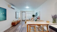 This thoughtfully designed single-floor residence offers a seamless blend of modern aesthetics and functional living. The open-concept layout connects the living, dining, and kitchen areas, creating a bright and cohesive space bathed in natural light from large windows fitted with white blinds. Wide-plank wood-look flooring runs throughout, lending warmth and continuity to every room.

The living area is generously proportioned, featuring a wall-mounted mini-split HVAC unit for individualized climate control and ample wall space accented by bold, colorful artwork. A glass-panel door provides access to the exterior, further enhancing the flow of natural light.

The kitchen is equipped with stainless steel appliances, including a range and over-the-range microwave, complemented by two-tone cabinetry — white upper cabinets paired with wood-finish lower drawers — and clean white countertops that offer practical workspace.

The bedroom is a tranquil retreat with neutral walls, a bright window, a wall-mounted mini-split unit, and access to a dedicated walk-in closet area with built-in hanging rods. The full bathroom features a tub-shower combination with white subway tile surround, wood-look flooring, and a clean, minimalist design that connects directly to the bedroom for added convenience.