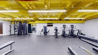 This 990-square-foot exercise room in Columbia, MD, is a testament to modern fitness design. The space is characterized by its striking yellow ceiling, which creates a vibrant and energizing atmosphere. Fluorescent lighting fixtures are strategically placed to ensure excellent visibility throughout the area.

The room is equipped with a comprehensive array of exercise equipment, catering to various workout preferences. Cardio machines, including treadmills and ellipticals, are positioned along the walls. Strength training equipment, such as weight machines and free weights, are distributed throughout the space, allowing for a well-rounded fitness routine.

The floor is covered with durable, dark gray material, likely rubber or specialized gym flooring, providing comfort and safety during workouts. White painted brick walls contrast nicely with the yellow ceiling, creating a clean and bright environment.

Storage solutions are integrated into the design, with equipment racks and possibly lockers visible. The layout appears spacious and well-organized, allowing for easy navigation between different workout zones. High ceilings contribute to a sense of openness, potentially improving air circulation.

This fitness facility combines functionality with a modern aesthetic, offering a welcoming space for exercise enthusiasts. Its design demonstrates a thoughtful approach to creating an environment that is both practical for workouts and visually stimulating.