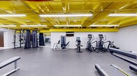 Located at 5764 Stevens Forest Rd in Columbia, MD, this fitness center offers a generously sized, single-floor exercise room designed to support a comprehensive workout experience. The space immediately stands out with its bold industrial aesthetic — a vivid yellow-painted exposed ceiling with structural beams, ductwork, and bright linear fluorescent lighting that illuminates the entire floor with excellent visibility.

The exercise room is thoughtfully organized into distinct training zones. The cardio area is outfitted with multiple treadmills, elliptical trainers, a recumbent bike, and a rowing machine, all featuring modern digital displays. The strength training section includes a variety of Matrix-branded resistance machines, a leg press, and a dedicated free weights zone with a full dumbbell rack and adjustable benches. Mirrored walls in the strength area enhance the sense of space and allow users to monitor their form during workouts.

Decorative wood-clad columns add a warm contrast to the otherwise industrial palette, while the durable rubber flooring throughout provides both safety and comfort. Additional amenities include a wall-mounted television, stability balls, resistance accessories, and ample open floor space for functional training. The layout is efficient and accessible, making this fitness center a versatile facility for a wide range of exercise routines.