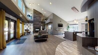 Located at 151 S Joliet Cir in Aurora, Colorado, this single-story community clubhouse delivers a sleek, contemporary design with an open, flowing layout. The space immediately impresses with its soaring vaulted ceilings that amplify the sense of openness throughout. The central lounge area serves as the heart of the space, anchored by a striking floor-to-ceiling tiled fireplace surround finished in rich, striated gray tones. Comfortable sectional seating, a round coffee table, and curated wall art create a welcoming and sophisticated atmosphere. Wide-plank wood-look flooring flows seamlessly throughout, unifying the open floor plan. Adjacent to the lounge, a full community kitchen features bold chartreuse cabinetry, a mosaic tile backsplash, stainless steel appliances, and a large dark granite island with bar seating, illuminated by modern pendant lighting. Large windows flood this area with abundant natural light and views of the surrounding exterior. The leasing office area offers an open workspace with multiple desks, large windows overlooking the outdoor grounds, and high ceilings that maintain the airy feel consistent with the rest of the space. Stone accent stools and tile flooring add textural contrast in the entry zone. Throughout, the design balances modern elegance with functional, community-focused amenities.