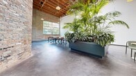 Located at 4100 Commerce St, this thoughtfully renovated single-floor industrial space blends raw architectural character with modern, biophilic design. The open-plan layout offers a generous, flexible footprint flooded with natural light through large steel-framed windows that frame views of surrounding greenery. Exposed brick walls run the length of the space, complemented by soaring wood-beam ceilings with visible structural rafters and industrial ductwork that add warmth and texture overhead. Polished concrete floors anchor the space with a sleek, durable finish that reflects light throughout. A standout feature is the dramatic large-scale planter housing lush tropical palms and foliage, serving as a natural room divider between a collaborative workstation area and a casual lounge. The workstation zone is equipped with a long communal table and ergonomic rolling chairs, while the lounge area features round bistro-style tables with modern green seating. Pendant lighting hangs throughout, adding ambient warmth to the industrial aesthetic. A set of steel-framed glass doors and an entry vestibule with exposed brick provide a distinct sense of arrival. The overall ambiance is open, airy, and vibrant — balancing industrial heritage with a fresh, nature-inspired atmosphere ideal for creative or professional use.