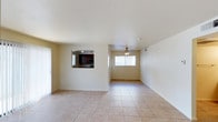 This single-story home offers a clean, functional layout designed for comfortable everyday living. The main living area is finished with large, light-toned ceramic tile flooring that extends throughout, lending a cohesive and easy-to-maintain aesthetic to the space. Neutral walls and a white textured ceiling contribute to an airy, bright atmosphere, while a ceiling fan with a light fixture provides both ventilation and overhead illumination.

Natural light fills the living area through a window fitted with white blinds, keeping the space feeling open and welcoming. A distinctive pass-through window cut into the wall connects the living area to the kitchen, offering a practical and social design feature. The kitchen, visible through the pass-through, showcases rich dark cabinetry paired with stainless steel appliances and recessed lighting, creating a functional cooking space with a warm contrast to the lighter tones of the main living area.

The overall layout is straightforward and versatile, providing an open floor plan that allows for flexible furniture arrangements. The home's simple, well-maintained finishes make it a practical and inviting space suitable for a variety of occupants.