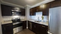 This single-floor residence offers one bedroom and one bathroom, designed with a focus on modern aesthetics and functional living. The kitchen serves as a standout feature, showcasing rich dark-espresso cabinetry that extends to the upper and lower walls, providing generous storage throughout. Light granite countertops create a striking contrast against the dark cabinetry, while a classic white subway tile backsplash adds a clean, timeless backdrop to the cooking area. Stainless steel appliances, including a range with an over-the-range microwave and a dishwasher, lend a sleek, contemporary feel to the space. A stainless steel undermount sink with a modern arc-neck faucet is positioned beneath ample counter space, making the kitchen both practical and visually cohesive. Crown molding along the ceiling adds an elegant architectural detail, elevating the overall finish of the space. The warm-toned hardwood-style flooring grounds the room and complements the cabinetry, creating a unified, polished look throughout this well-designed residence.