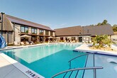 Located at 4660 Derrick Rd in Union City, Georgia, this outdoor amenity space centers around a generously sized resort-style swimming pool with crystal-clear blue water. The pool features a gradual depth progression, in-water lounge chairs, and stainless steel entry rails, making it both functional and inviting. Surrounding the pool is a spacious concrete sundeck lined with cushioned lounge chairs and market umbrellas, providing ample space for relaxation under the Georgia sun. Tropical plantings, including palm trees and ornamental shrubs, are integrated throughout the pool surround, adding a lush, resort-like atmosphere. The adjacent clubhouse building is a visual centerpiece, showcasing a blend of stone veneer, horizontal wood siding, and large floor-to-ceiling windows topped with a pitched metal roof — a design that balances modern and traditional architectural elements. Multi-story residential buildings with covered balconies frame the perimeter of the pool area, creating an enclosed and community-oriented outdoor environment. Mature trees border the property, providing a natural backdrop and shade. The overall ambiance is that of an upscale, amenity-rich residential community designed for comfort and outdoor living.
