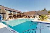 Located at 4660 Derrick Rd in Union City, Georgia, this outdoor amenity space centers around a generously sized resort-style swimming pool with crystal-clear blue water. The pool features a gradual depth progression, in-water lounge chairs, and stainless steel entry rails, making it both functional and inviting. Surrounding the pool is a spacious concrete sundeck lined with cushioned lounge chairs and market umbrellas, providing ample space for relaxation under the Georgia sun. Tropical plantings, including palm trees and ornamental shrubs, are integrated throughout the pool surround, adding a lush, resort-like atmosphere. The adjacent clubhouse building is a visual centerpiece, showcasing a blend of stone veneer, horizontal wood siding, and large floor-to-ceiling windows topped with a pitched metal roof — a design that balances modern and traditional architectural elements. Multi-story residential buildings with covered balconies frame the perimeter of the pool area, creating an enclosed and community-oriented outdoor environment. Mature trees border the property, providing a natural backdrop and shade. The overall ambiance is that of an upscale, amenity-rich residential community designed for comfort and outdoor living.