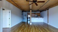 This single-floor loft showcases a compelling blend of industrial and rustic design elements, creating a warm yet contemporary atmosphere. The open-concept living area is anchored by rich, polished hardwood floors that reflect natural and ambient light beautifully throughout the space. Overhead, exposed wooden plank ceilings with visible structural beams lend a raw, authentic character, while ceiling fans provide both function and style. Structural columns run from floor to ceiling, adding to the loft's industrial charm without interrupting the generous sense of openness. The kitchen is thoughtfully integrated into the open layout, featuring warm wood cabinetry, stainless steel appliances including a refrigerator and microwave, and a breakfast bar with an overhang that offers casual seating. Track lighting and pendant fixtures are strategically placed throughout, ensuring the space is well-lit while complementing the overall aesthetic. Exposed utility conduits along the ceiling further reinforce the building's converted industrial heritage. The expansive, unobstructed floor plan offers exceptional flexibility for living and entertaining, making this loft a distinctive and versatile space.