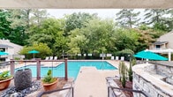 Located at 7303 Bryn Athyn Way in Raleigh, North Carolina, this outdoor amenity space centers around a generously sized swimming pool with clear, blue-green water. The pool deck is finished with a smooth concrete surround and offers ample space for relaxation, furnished with lounge chairs and tables shaded by vibrant teal umbrellas. The area is thoughtfully enclosed by a sturdy black iron fence, providing both security and an open, airy feel while framing views of the mature trees and dense greenery that border the property on multiple sides.

A clubhouse building anchors one end of the pool area, featuring a welcoming entrance with wide stone steps, metal handrails, and a bold set of blue French doors framed by white columns. A raised deck with stone accents adjoins the building, offering an elevated outdoor seating area with additional patio furniture — ideal for gatherings or casual outdoor dining. A storage shed sits conveniently nearby. The surrounding landscape is rich with tall trees and lush shrubs, creating a private, natural backdrop that enhances the overall ambiance of the space. The combination of well-maintained hardscaping, thoughtful landscaping, and functional amenities makes this a comfortable and inviting outdoor destination.