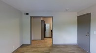 This single-floor space offers a clean, functional layout designed with simplicity and practicality in mind. The main living area features wood-look plank flooring that runs seamlessly throughout, providing a warm and cohesive foundation underfoot. Neutral white walls create a bright, open atmosphere, while gray trim detailing around the doorways adds a subtle modern contrast.

A wide doorway opening connects the living area to the kitchen, promoting an easy flow between spaces. The kitchen is equipped with a full-size refrigerator and additional appliances, set against dark cabinetry that creates a striking visual contrast with the lighter surroundings. The flooring continues consistently from room to room, reinforcing the unified feel of the layout.

Overhead, a ceiling-mounted HVAC vent ensures climate control throughout the space, and a smoke detector is visibly installed, reflecting attention to safety. The overall design is minimal and move-in ready, with a neutral palette that provides a versatile backdrop for a variety of interior styles. The space is well-suited for those seeking a straightforward, low-maintenance environment with a modern aesthetic sensibility.
