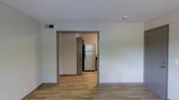 This single-floor space offers a clean, functional layout designed with simplicity and practicality in mind. The main living area features wood-look plank flooring that runs seamlessly throughout, providing a warm and cohesive foundation underfoot. Neutral white walls create a bright, open atmosphere, while gray trim detailing around the doorways adds a subtle modern contrast.

A wide doorway opening connects the living area to the kitchen, promoting an easy flow between spaces. The kitchen is equipped with a full-size refrigerator and additional appliances, set against dark cabinetry that creates a striking visual contrast with the lighter surroundings. The flooring continues consistently from room to room, reinforcing the unified feel of the layout.

Overhead, a ceiling-mounted HVAC vent ensures climate control throughout the space, and a smoke detector is visibly installed, reflecting attention to safety. The overall design is minimal and move-in ready, with a neutral palette that provides a versatile backdrop for a variety of interior styles. The space is well-suited for those seeking a straightforward, low-maintenance environment with a modern aesthetic sensibility.