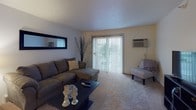 This single-floor unit in Angola, Indiana, offers a well-organized layout designed for comfortable everyday living. The living room serves as the central gathering space, featuring plush carpeting, a sectional sofa, and a sliding glass door framed by vertical blinds that opens to an outdoor view and brings in natural light. A wall-mounted air conditioning unit ensures year-round comfort.

Adjacent to the living area, a compact dining space accommodates a bistro-style table and chairs beneath a ceiling light fixture, situated near the entry door. The galley-style kitchen is efficiently arranged with white cabinetry accented by warm wood trim, a double sink, full-sized appliances including a refrigerator and dishwasher, and in-unit laundry. Light flooring runs throughout the kitchen, keeping the space bright.

The primary bedroom is carpeted and features a ceiling fan with light, a window with outdoor views, and a closet accessible through a wood-paneled door. A second carpeted room with two closets and a window serves well as a home office or additional bedroom. The full bathroom includes a tub-shower combination, tile flooring, a vanity with countertop, and baseboard heating.