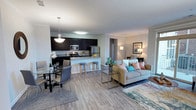 This single-floor residence offers a thoughtfully designed open-concept layout that seamlessly integrates the kitchen, dining, and living areas into one cohesive, functional space. The kitchen is a standout feature, boasting rich dark cabinetry, granite countertops, stainless steel appliances, and a peninsula island with bar seating — ideal for casual dining or entertaining. Pendant lighting above the island and dining area adds a polished touch to the space.

The dining area accommodates a round glass-top table with upholstered chairs, accented by a decorative circular mirror that adds depth and character to the room. The living area is open and inviting, featuring durable wood-style plank flooring that extends throughout the main living space, providing warmth and a cohesive aesthetic.

Crown molding details along the ceilings lend an elevated architectural quality to the interior. Natural and ambient lighting work together to keep the space feeling bright and airy. A hallway leads to additional rooms, including what appears to be a well-lit bathroom, reinforcing the practical and comfortable nature of the floor plan. Overall, the space combines contemporary design elements with a warm, livable ambiance.