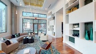 This thoughtfully designed single-floor communal space combines modern architecture with refined interior details to create an inviting and functional environment. The centerpiece is a grand lounge area defined by soaring double-height ceilings and expansive floor-to-ceiling windows framed in sleek black metal, flooding the space with natural light and offering views of lush outdoor palm-lined surroundings. A striking cascade of pendant light fixtures hangs overhead, adding a sculptural focal point to the room. Rich hardwood flooring flows throughout, anchoring the space with warmth and continuity. The lounge is furnished with contemporary seating arrangements — including curved sofas, swivel chairs, and accent tables — arranged atop a vibrant patterned area rug, encouraging both relaxed gathering and casual conversation. A dramatic floor-to-ceiling built-in shelving unit lines one wall, offering generous display space with an open, airy composition. A dedicated corridor features a row of glass-partitioned private rooms accessible via sleek glass-panel doors with dark frames, creating versatile enclosed spaces while maintaining visual openness. An additional seating alcove showcases a refined, minimalist aesthetic with textured plaster-effect walls in soft grey tones and large-format tile flooring, providing a quiet, contemplative retreat. Together, these distinct zones deliver a polished and cohesive amenity experience.