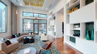 This thoughtfully designed single-floor communal space combines modern architecture with refined interior details to create an inviting and functional environment. The centerpiece is a grand lounge area defined by soaring double-height ceilings and expansive floor-to-ceiling windows framed in sleek black metal, flooding the space with natural light and offering views of lush outdoor palm-lined surroundings. A striking cascade of pendant light fixtures hangs overhead, adding a sculptural focal point to the room. Rich hardwood flooring flows throughout, anchoring the space with warmth and continuity. The lounge is furnished with contemporary seating arrangements — including curved sofas, swivel chairs, and accent tables — arranged atop a vibrant patterned area rug, encouraging both relaxed gathering and casual conversation. A dramatic floor-to-ceiling built-in shelving unit lines one wall, offering generous display space with an open, airy composition. A dedicated corridor features a row of glass-partitioned private rooms accessible via sleek glass-panel doors with dark frames, creating versatile enclosed spaces while maintaining visual openness. An additional seating alcove showcases a refined, minimalist aesthetic with textured plaster-effect walls in soft grey tones and large-format tile flooring, providing a quiet, contemplative retreat. Together, these distinct zones deliver a polished and cohesive amenity experience.