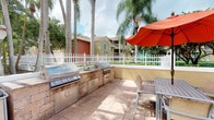 This inviting outdoor space is designed with entertaining and relaxation in mind, offering a thoughtfully equipped environment that evokes a resort-style atmosphere. The centerpiece of the area is a built-in outdoor kitchen, featuring dual stainless steel grills set into a sturdy stone-finished counter with integrated storage cabinets beneath. A utility sink positioned between the grills adds practicality, making this setup well-suited for hosting gatherings of any size. The patio surface is finished in warm-toned brick pavers that lend a classic, durable character to the space. Adjacent to the cooking station, a dining table paired with chairs sits beneath a large red market umbrella, offering a shaded spot to enjoy meals outdoors. The space is framed by tall, mature palm trees that provide natural privacy and a distinctly tropical ambiance. A white picket fence encloses the perimeter, giving the area a clean, well-defined boundary while maintaining an open and airy feel. The surrounding landscaping includes lush greenery that contributes to the overall sense of seclusion. The warm Florida-style architecture of the neighboring structures complements the outdoor aesthetic, completing a cohesive and welcoming environment.