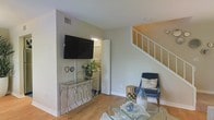This two-story home offers a practical and comfortable layout designed to maximize both functionality and livability. The main floor is anchored by a bright living area finished with warm hardwood-style flooring and neutral wall tones that create an inviting, versatile backdrop. A wall-mounted flat-screen TV is positioned above a sleek mirrored console table, while a glass coffee table and accent chair add stylish touches to the space. Natural light fills the room, highlighting the clean lines and open feel of the interior. A staircase with white railings leads to the upper floor, providing a clear visual connection between levels. Conveniently tucked just off the living area is an in-unit laundry closet, adding everyday practicality to the home. The upper floor extends the living space further, offering additional rooms that complement the well-organized floor plan below. Throughout the home, the design emphasizes a neutral, light-filled palette that lends itself to a range of décor styles. The combination of thoughtful storage solutions, multi-level living, and cohesive finishes makes this a highly functional and comfortable space suited for a variety of lifestyles.